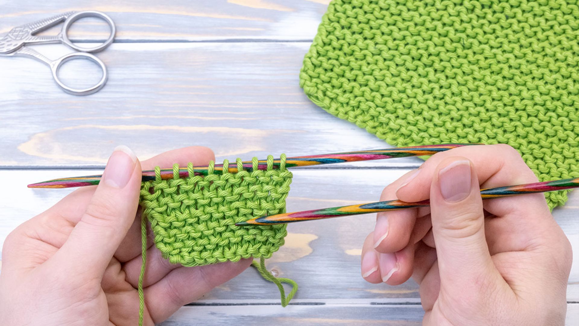 A person knitting a green piece of fabric with needles, focused on their craft.