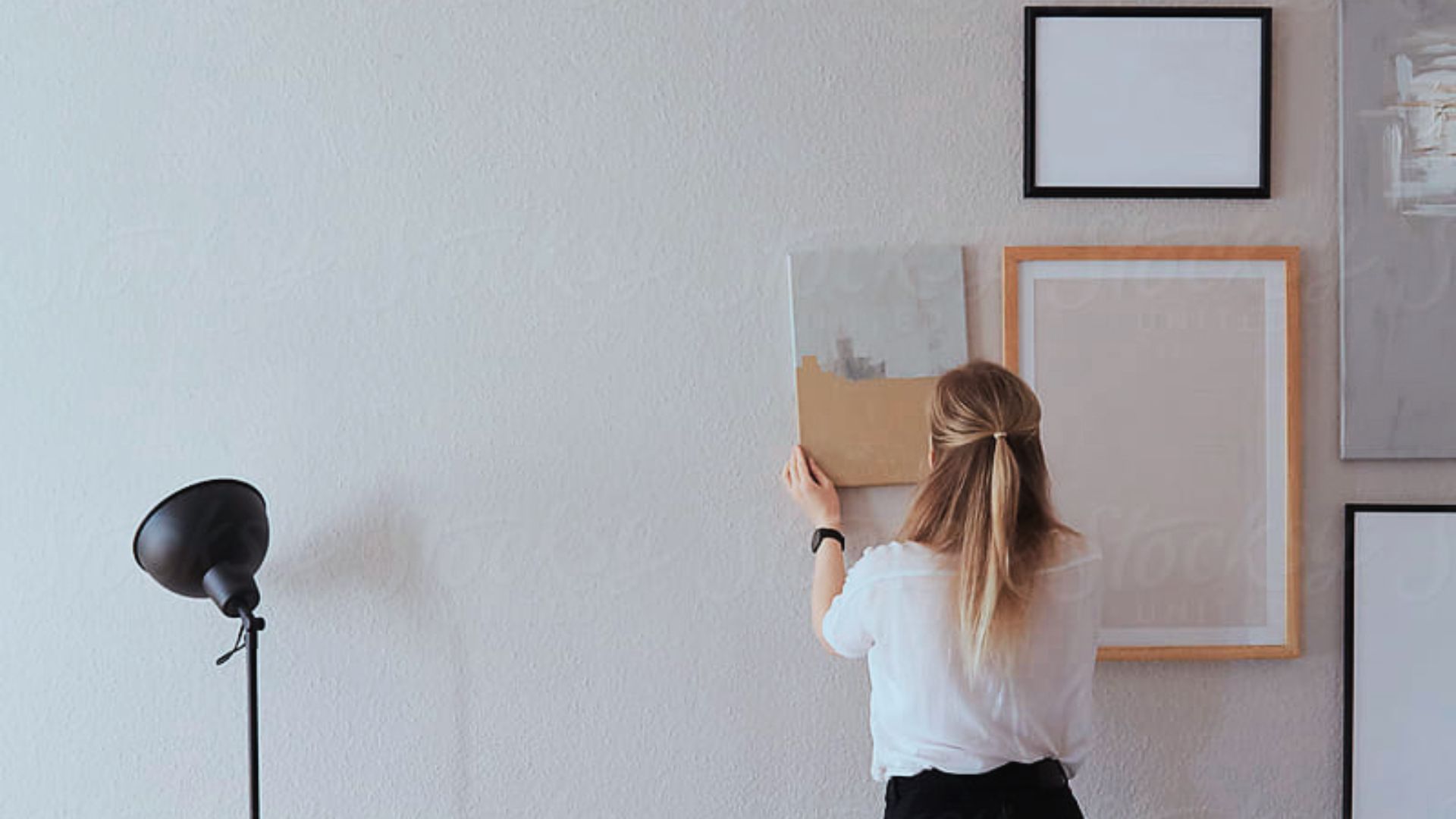 A woman hangs pictures on a wall beside a lamp, arranging her decor in a cozy living space.