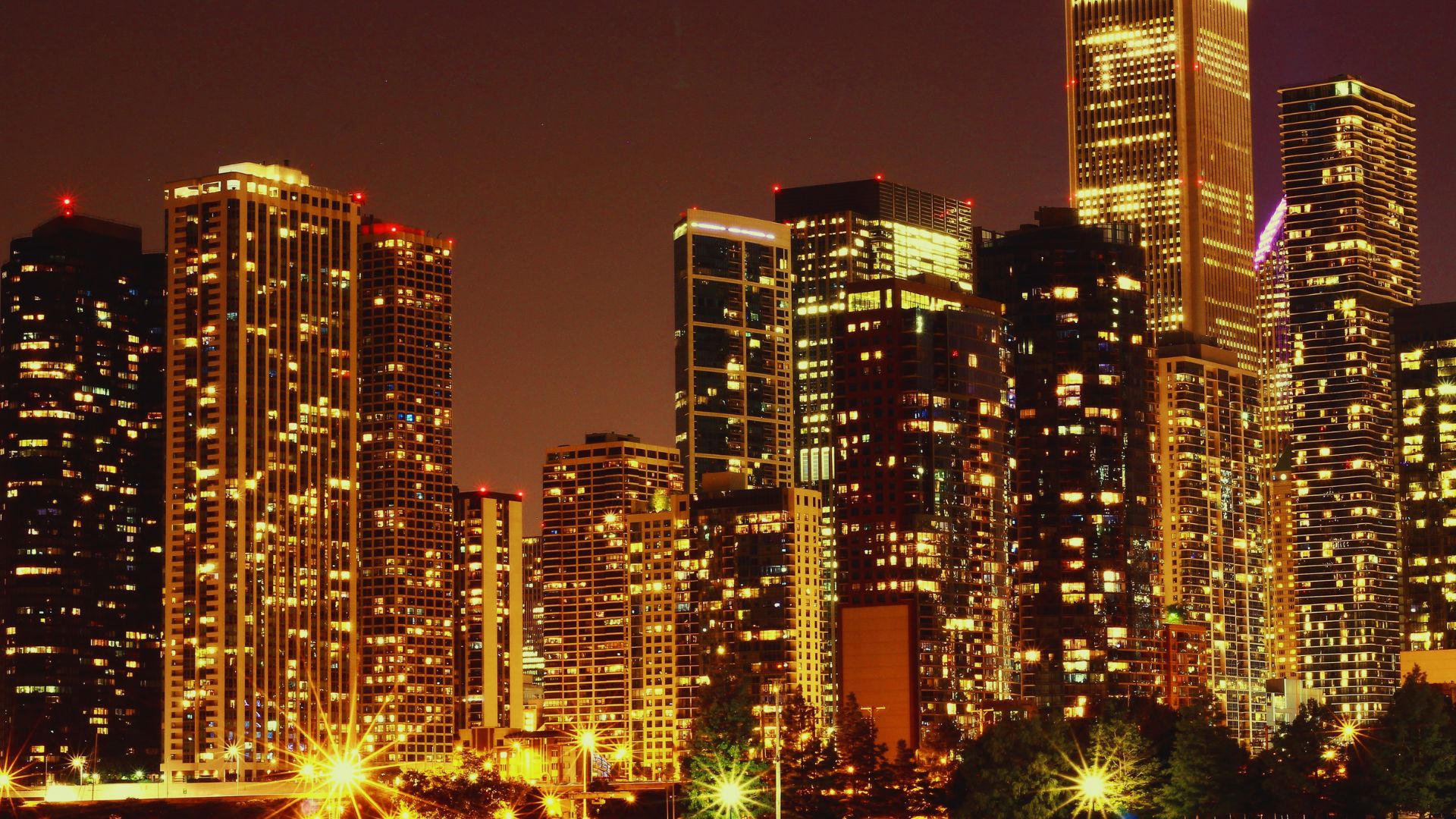 A vibrant cityscape at night featuring numerous tall buildings illuminated against the dark sky.