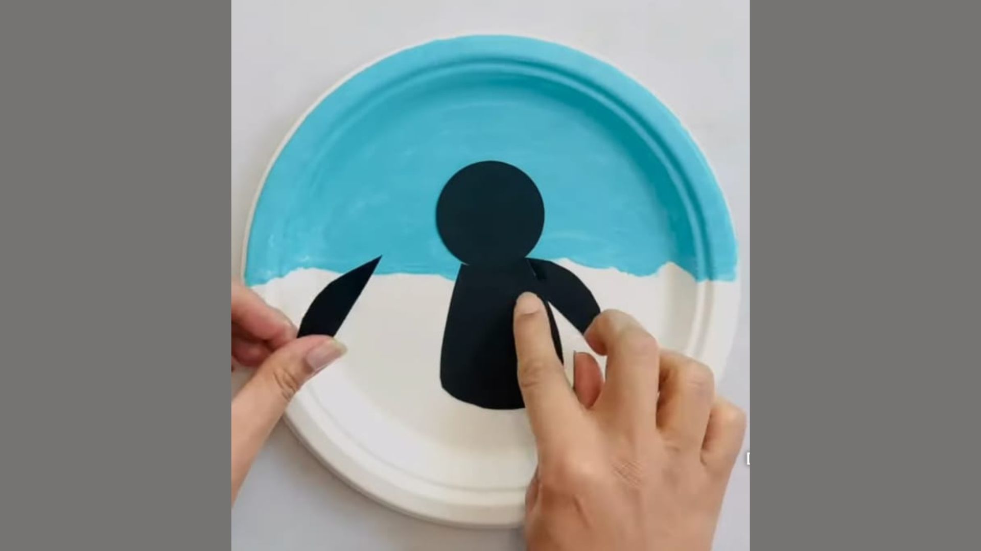A person paints a paper plate, creating a black and white penguin design on its surface.