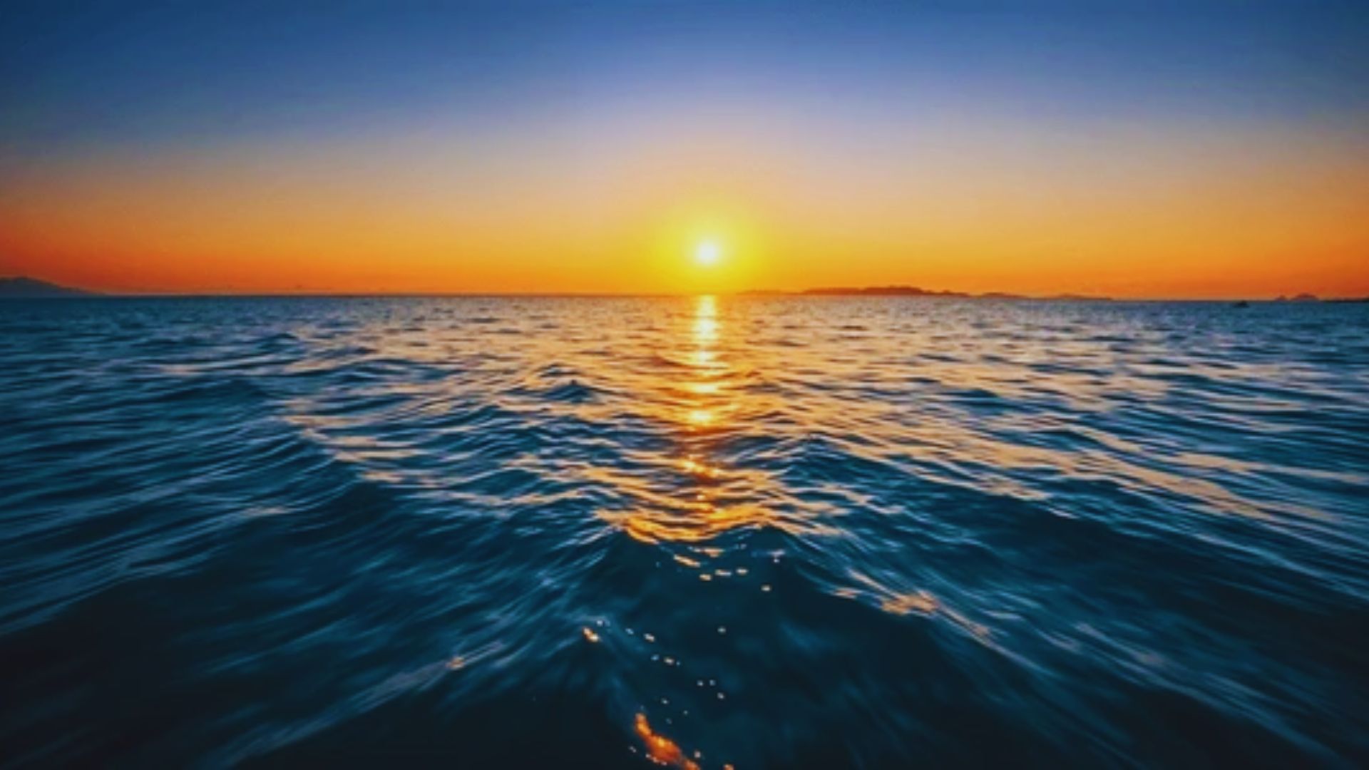 A vibrant sunset casts orange and pink hues over the calm ocean waves, creating a serene and picturesque scene.
