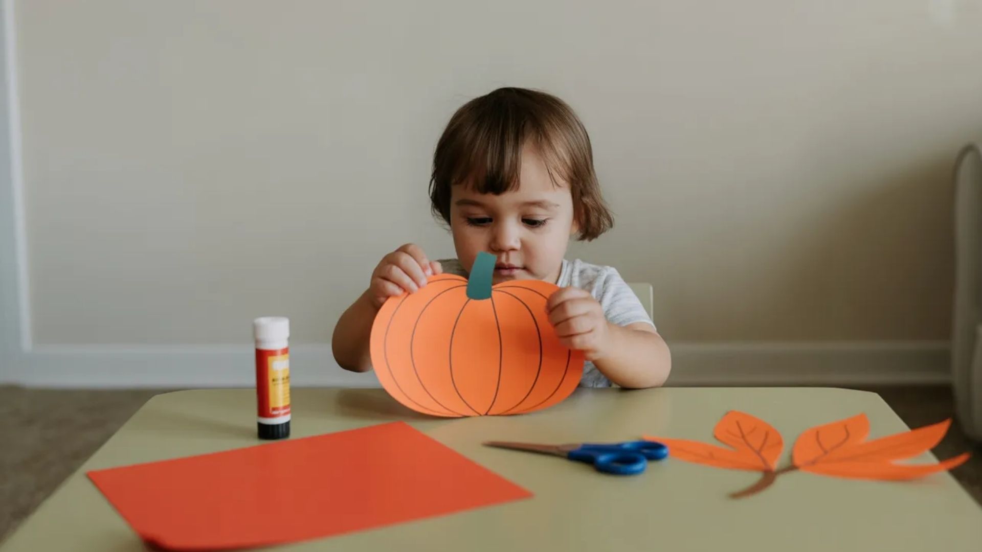 A toddler is engaged in creating a colorful pumpkin craft using various art supplies on a table.
