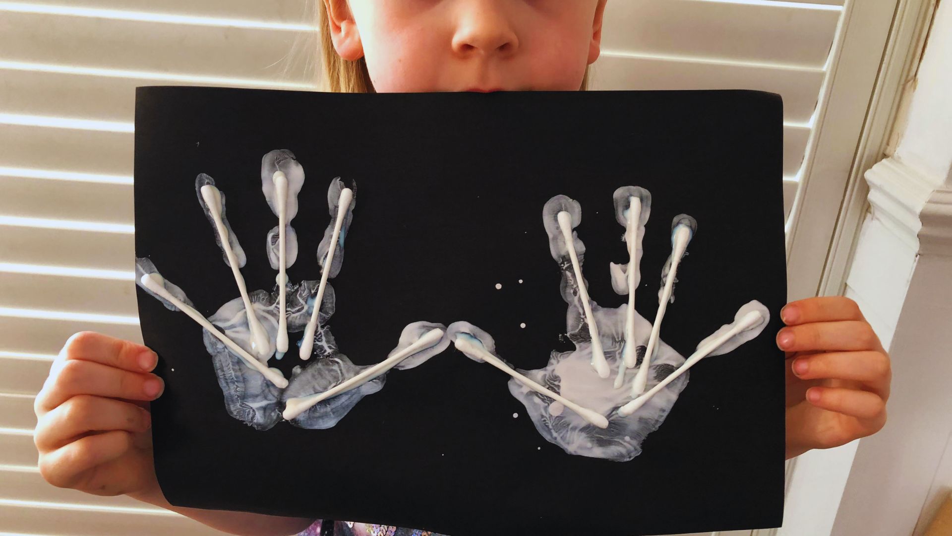 A young girl proudly displays a colorful handprint on paper, smiling at the camera.