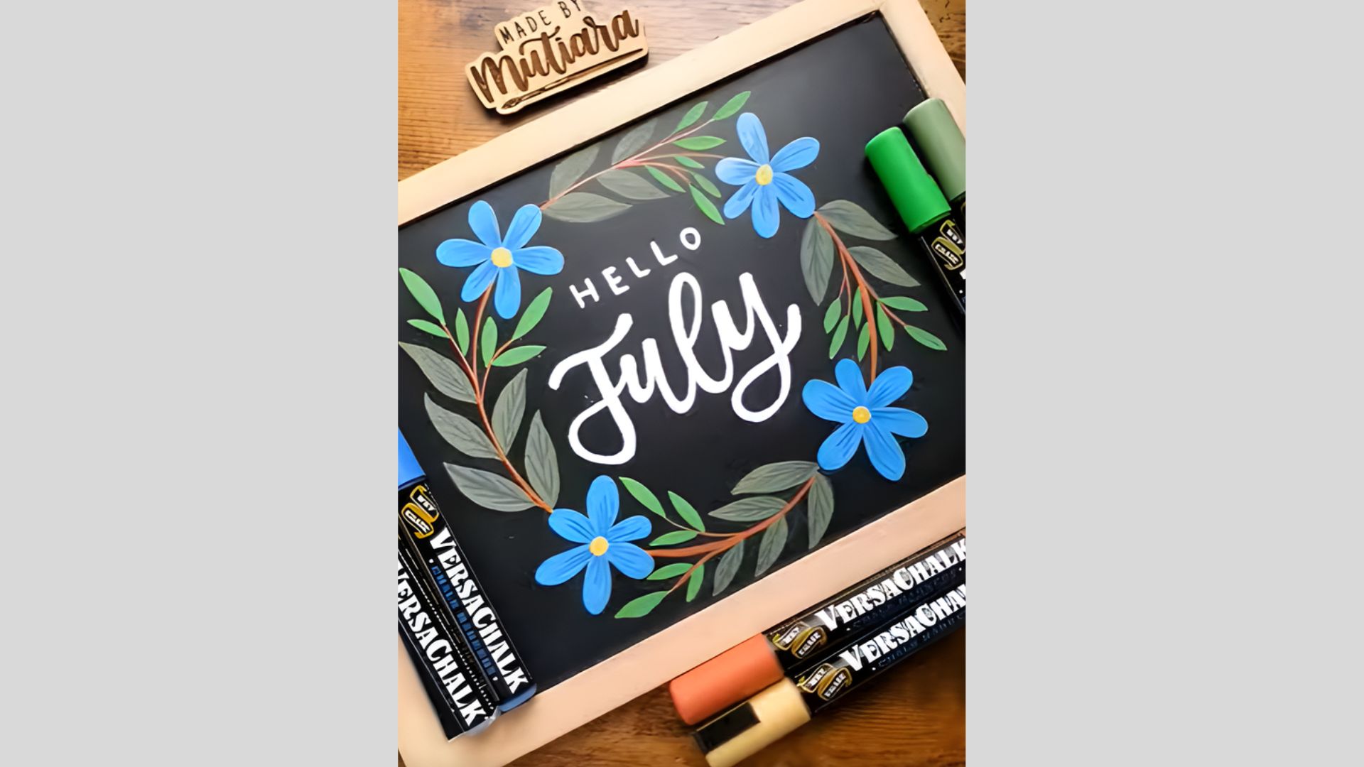A small framed chalkboard displaying “Hello July” in white lettering, surrounded by hand-drawn blue flowers and green leaves