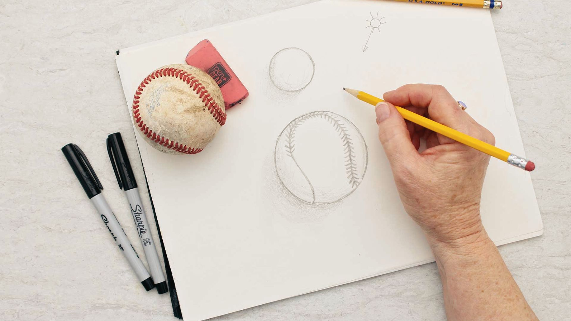 Person sketches realistic baseball with pencil and real ball as reference for easy art tutorial