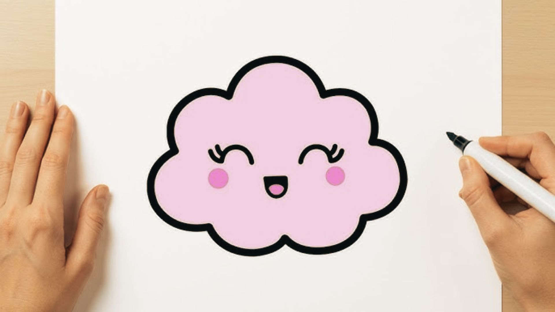 Top-down photo of hands drawing a pink kawaii cloud with big eyes and a happy face.