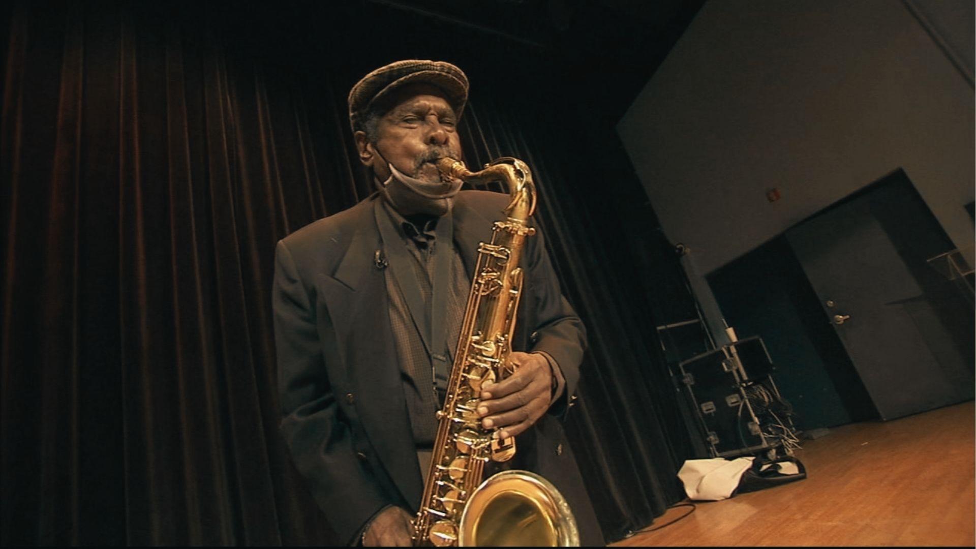 An elderly jazz musician in a flat cap and suit performs on stage with a golden tenor saxophone under warm lighting.