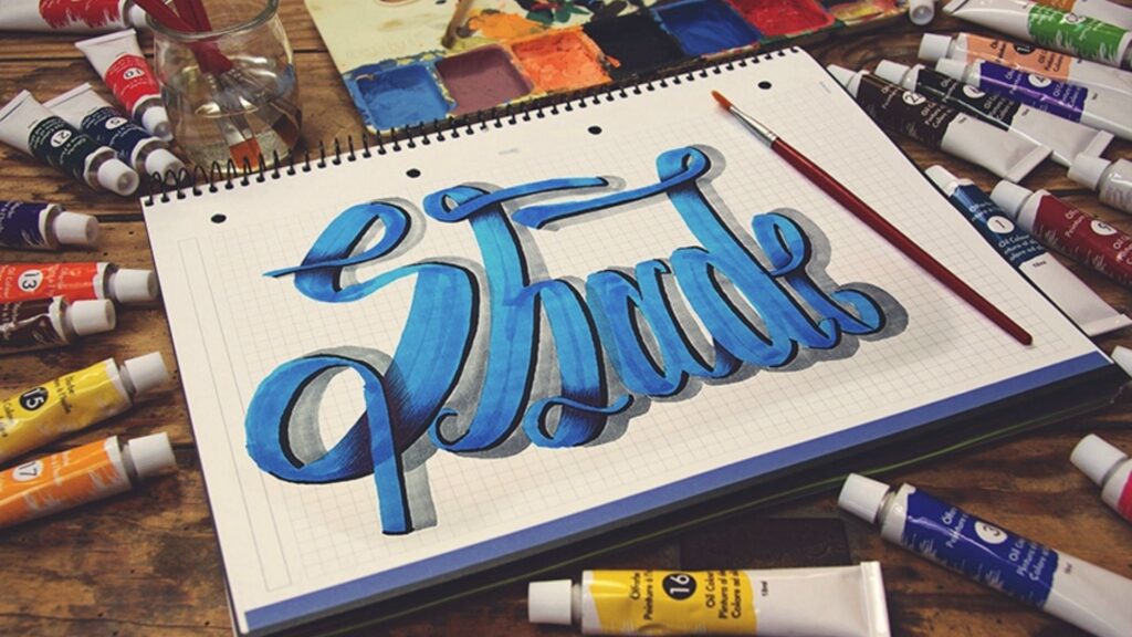Easy Shadow Letters: Step-by-Step Shading Tutorial The word “Shade” in blue brush lettering with gray drop shadow on a spiral grid sketchbook.