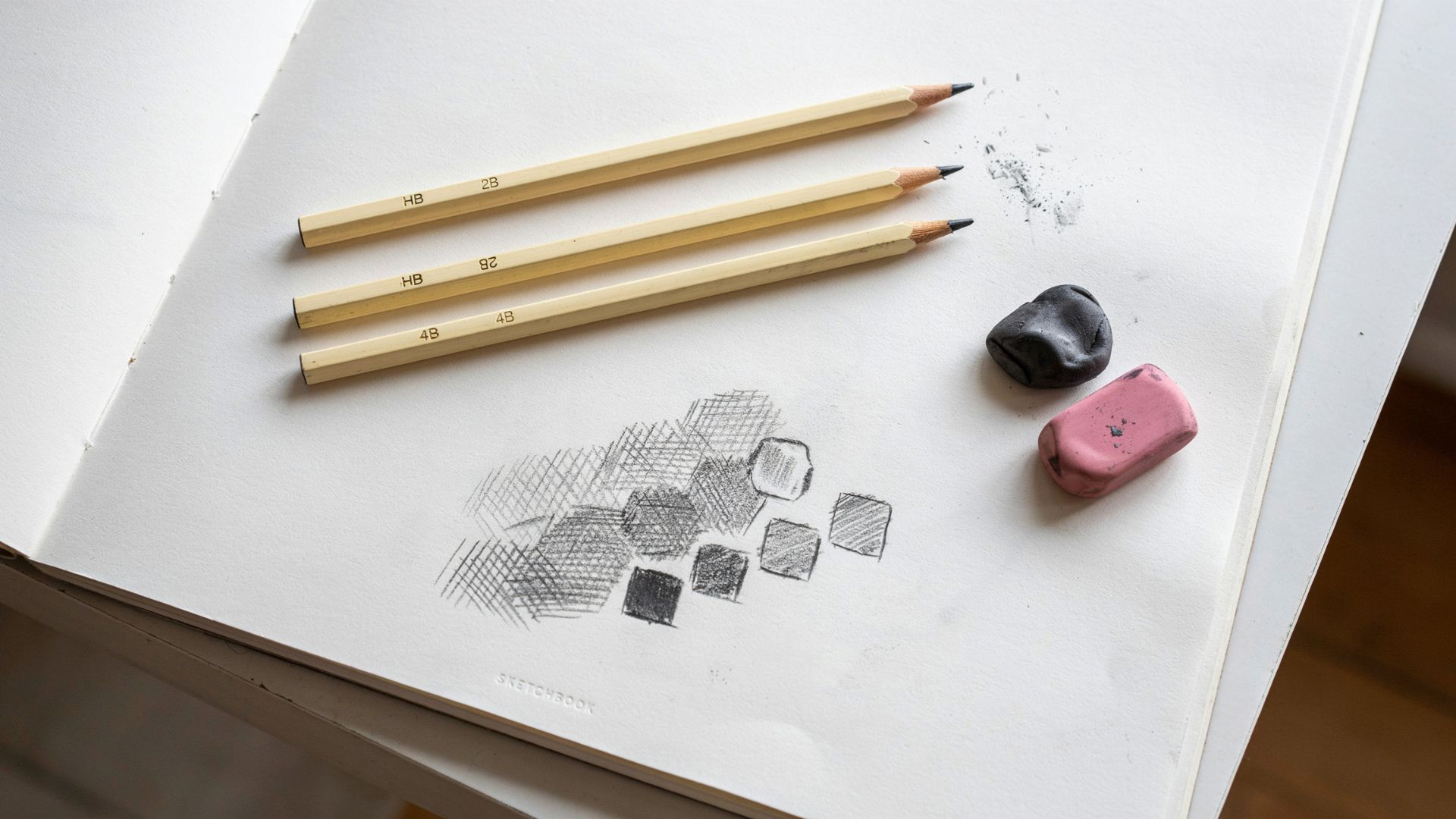 HB, 2B, and 4B pencils with a kneaded eraser and pink eraser displayed on a sketchbook showing shading and highlighting examples.