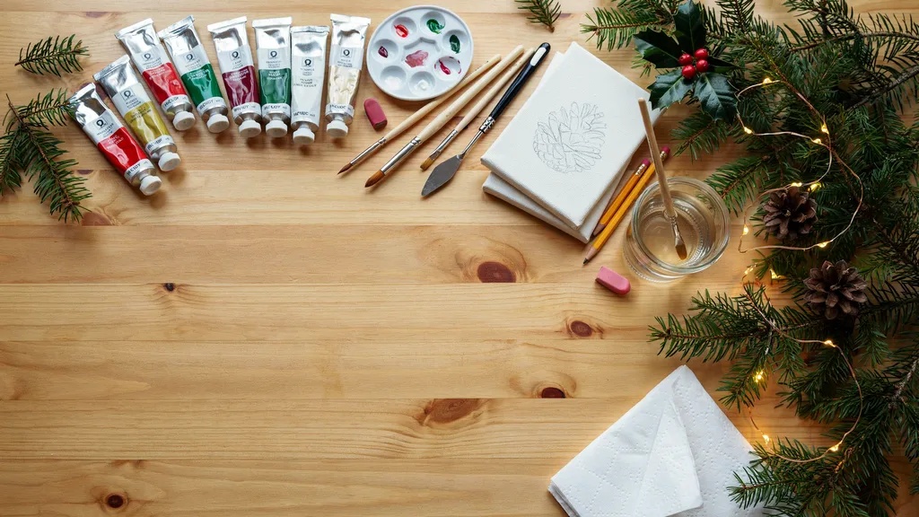 A top-down view of Christmas painting supplies including brushes, acrylic paints, palette, canvas sheets, pencils, water jar, and festive decorations arranged neatly.